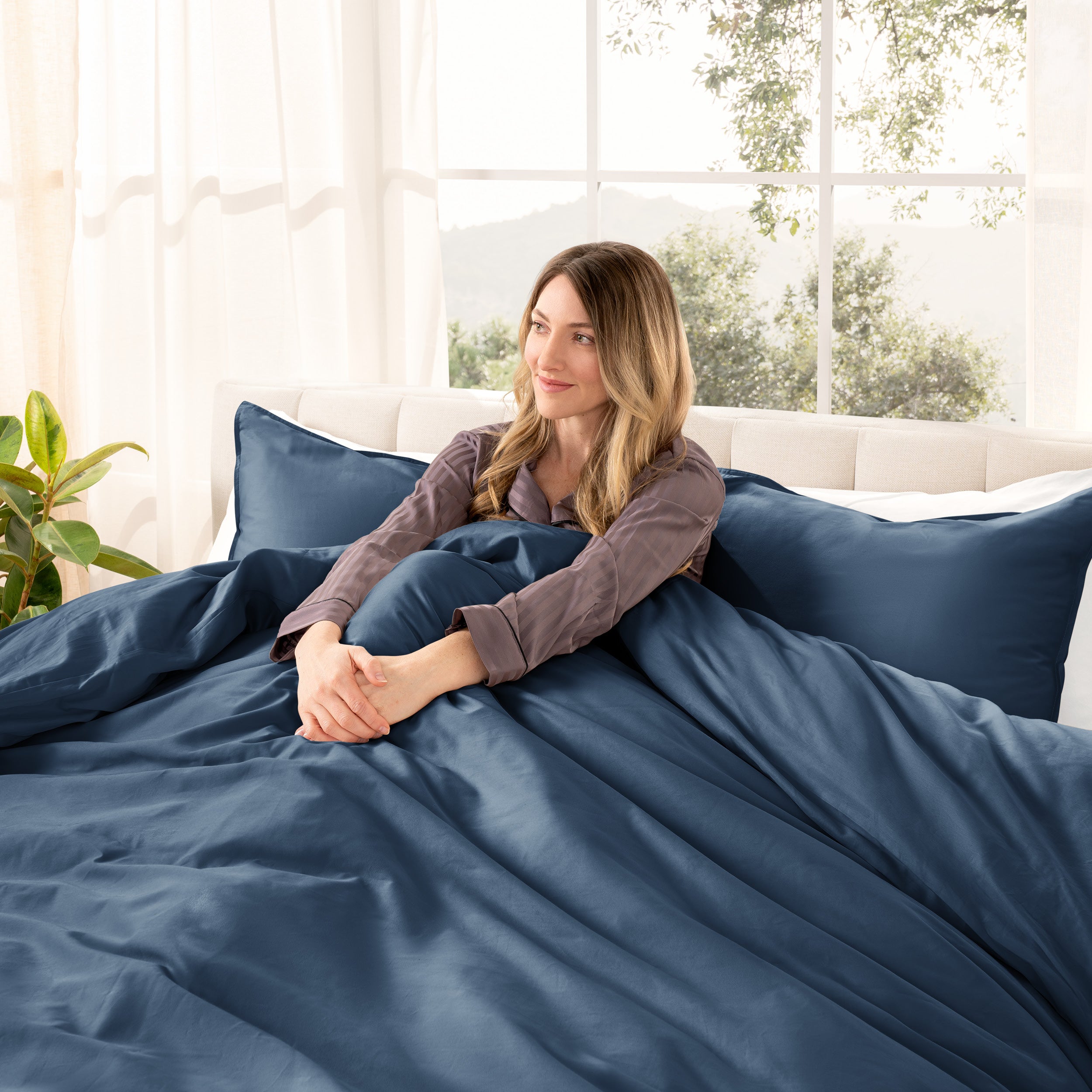 A woman sitting on a bed made with organic cotton sateen sheets and pillowcases