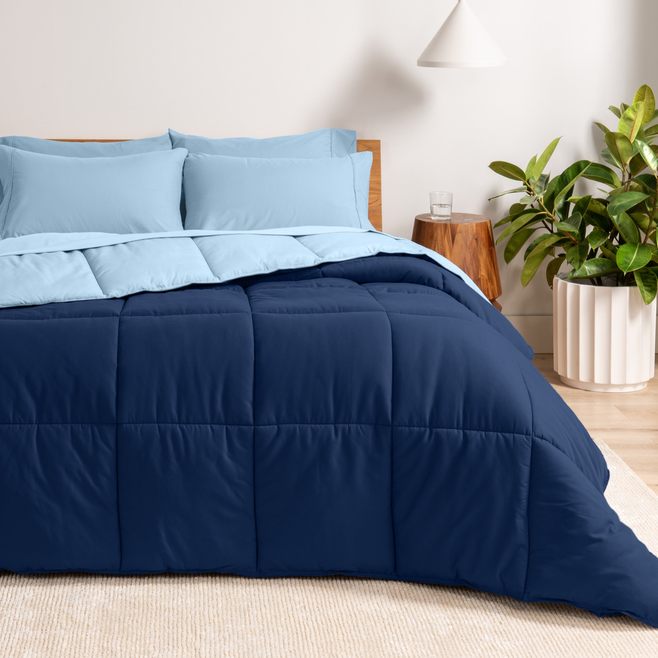 A wooden bed frame with a reversible comforter and sheet set on the mattress in a bright warm room.