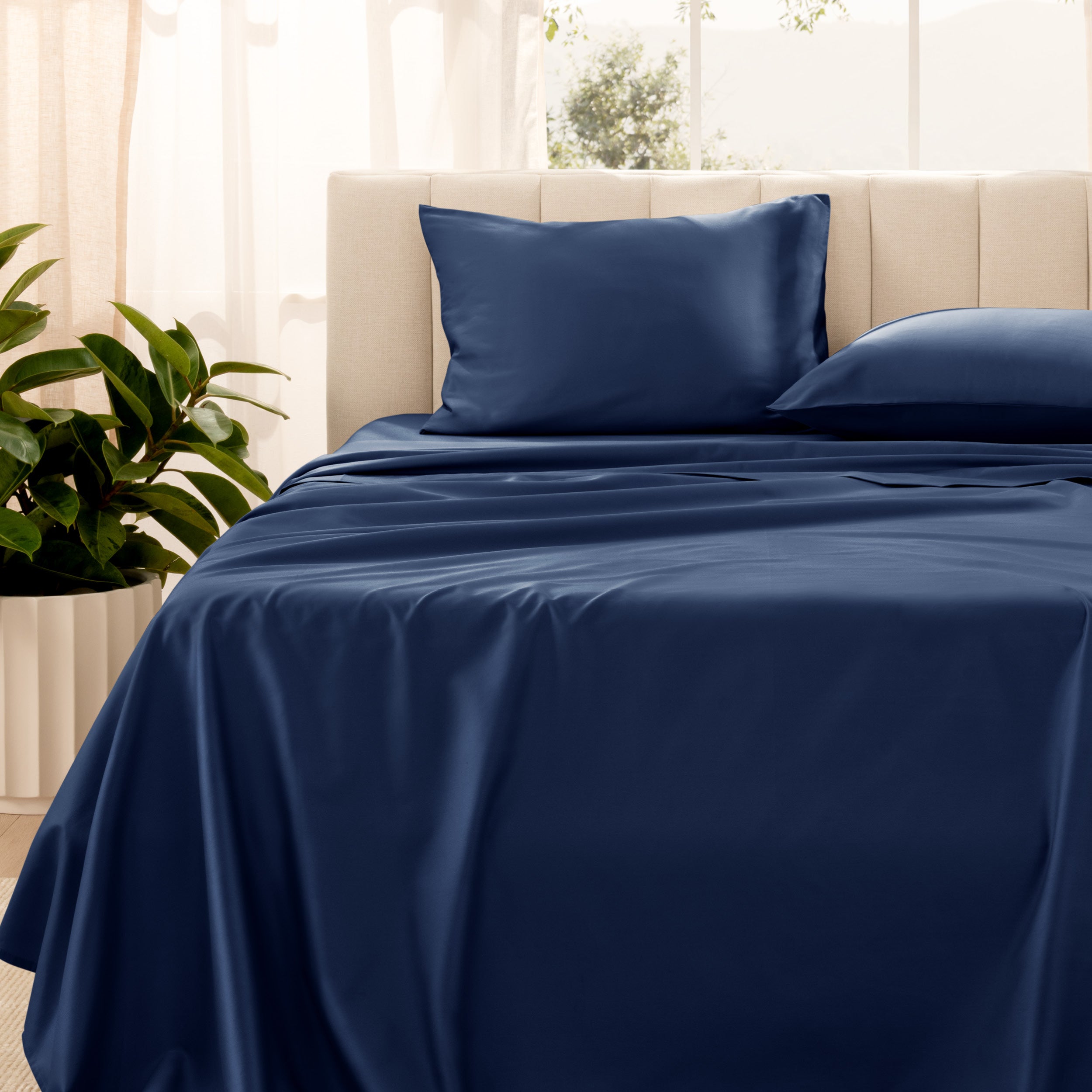 Organic sateen bedding on a bed with a beige headboard and plant in the corner.
