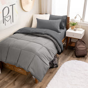 A wooden bed frame with a reversible comforter and sheet set on the mattress in a dorm style room.