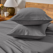 Two pillows with flannel pillowcases stacked on a bed with a wooden headboard and side table in the background.