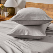 Two pillows with flannel pillowcases stacked on a bed with a wooden headboard and side table in the background.