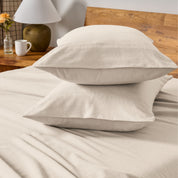 Two pillows with flannel pillowcases stacked on a bed with a wooden headboard and side table in the background.