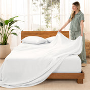 Woman arranging linen sheets on a wooden bed in a bright room with plants.