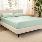 Organic cotton fitted sheet on a bed in a bedroom with a plant and window in the background