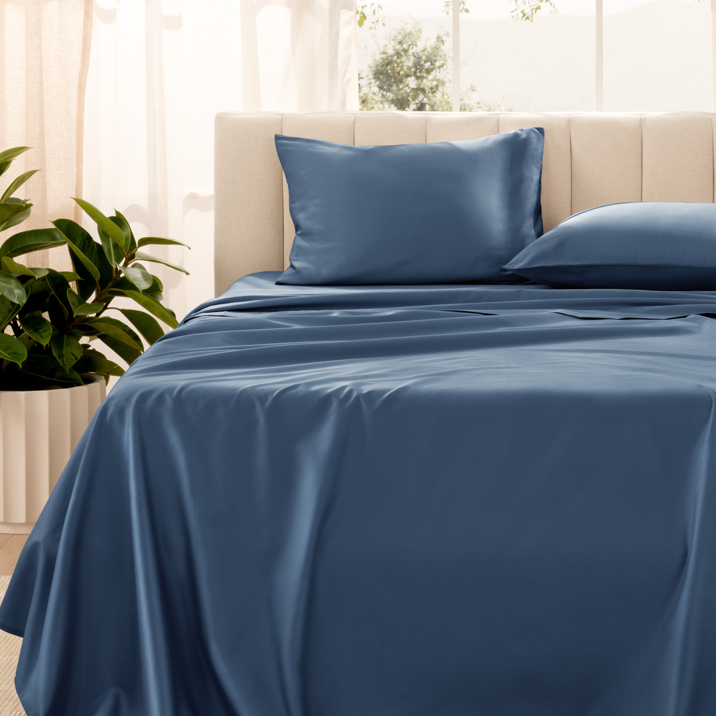 Organic sateen bedding on a bed with a beige headboard and plant in the corner.