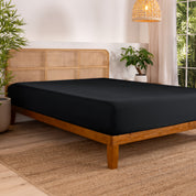 Bed with fitted sheet in a bedroom setting with plants and a lamp.