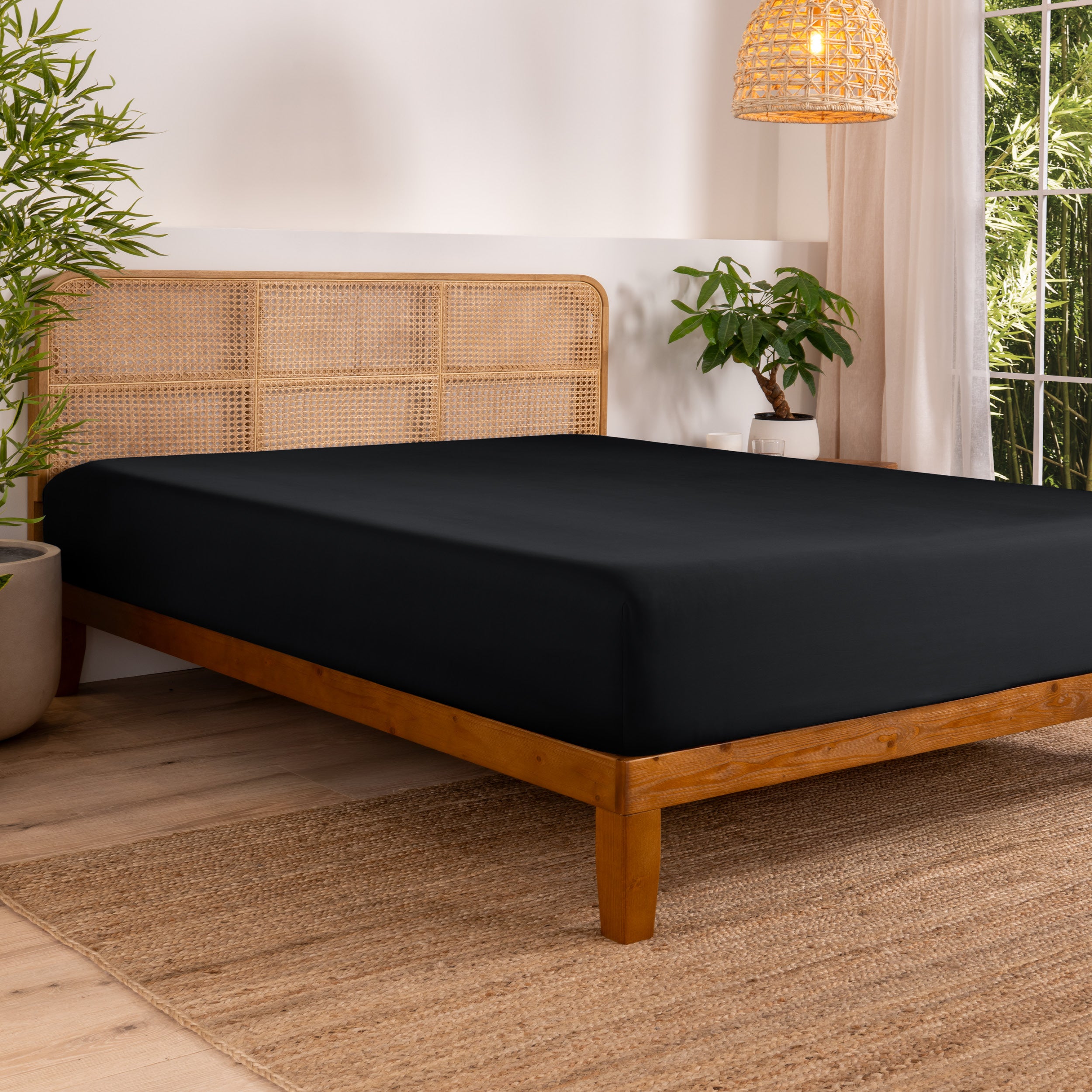 Bed with fitted sheet in a bedroom setting with plants and a lamp.