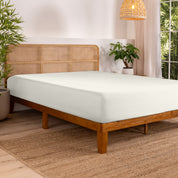 Bed with fitted sheet in a bedroom setting with plants and a lamp.