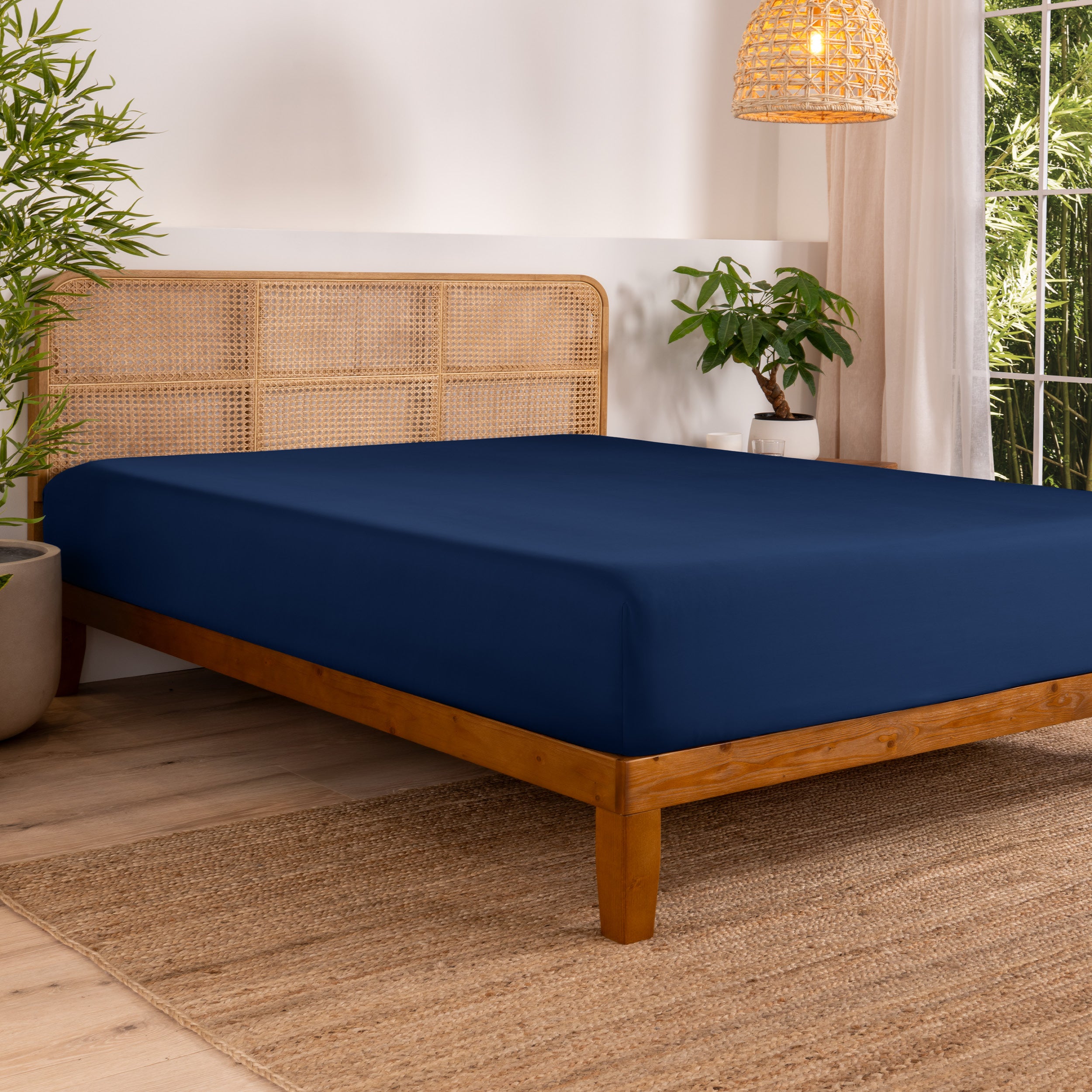 Bed with fitted sheet in a bedroom setting with plants and a lamp.