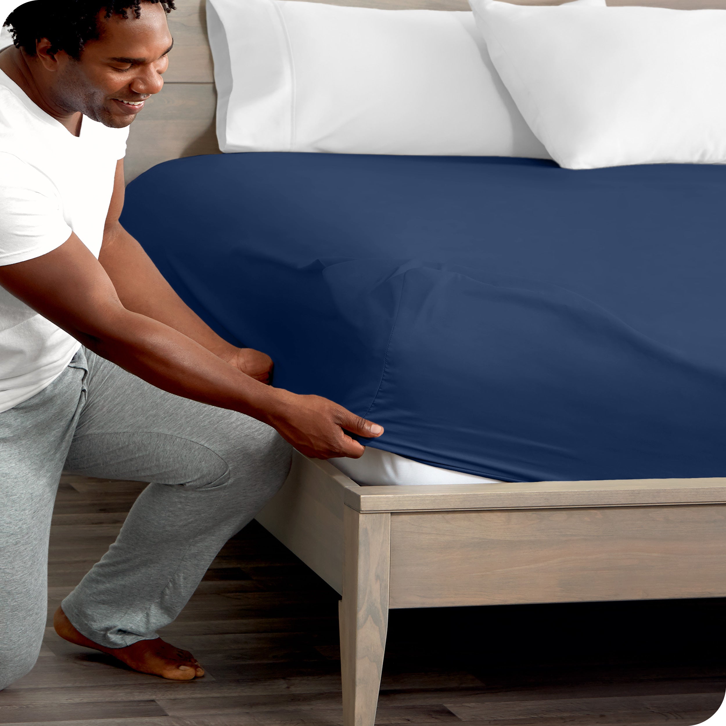 A man kneeling while putting a fitted sheet on a bed