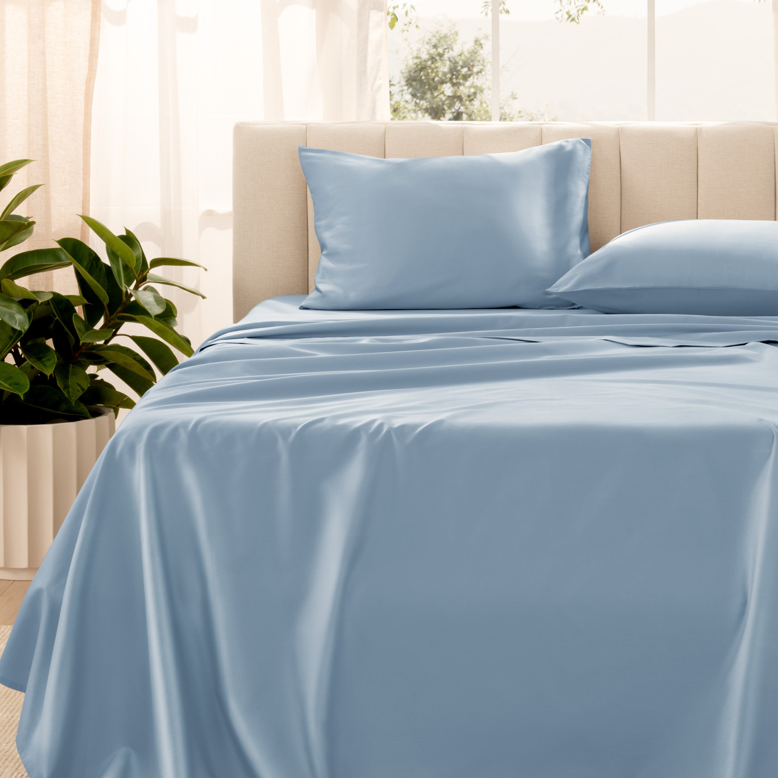 Organic sateen bedding on a bed with a beige headboard and plant in the corner.