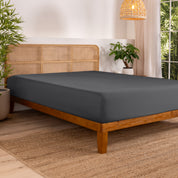 Bed with fitted sheet in a bedroom setting with plants and a lamp.