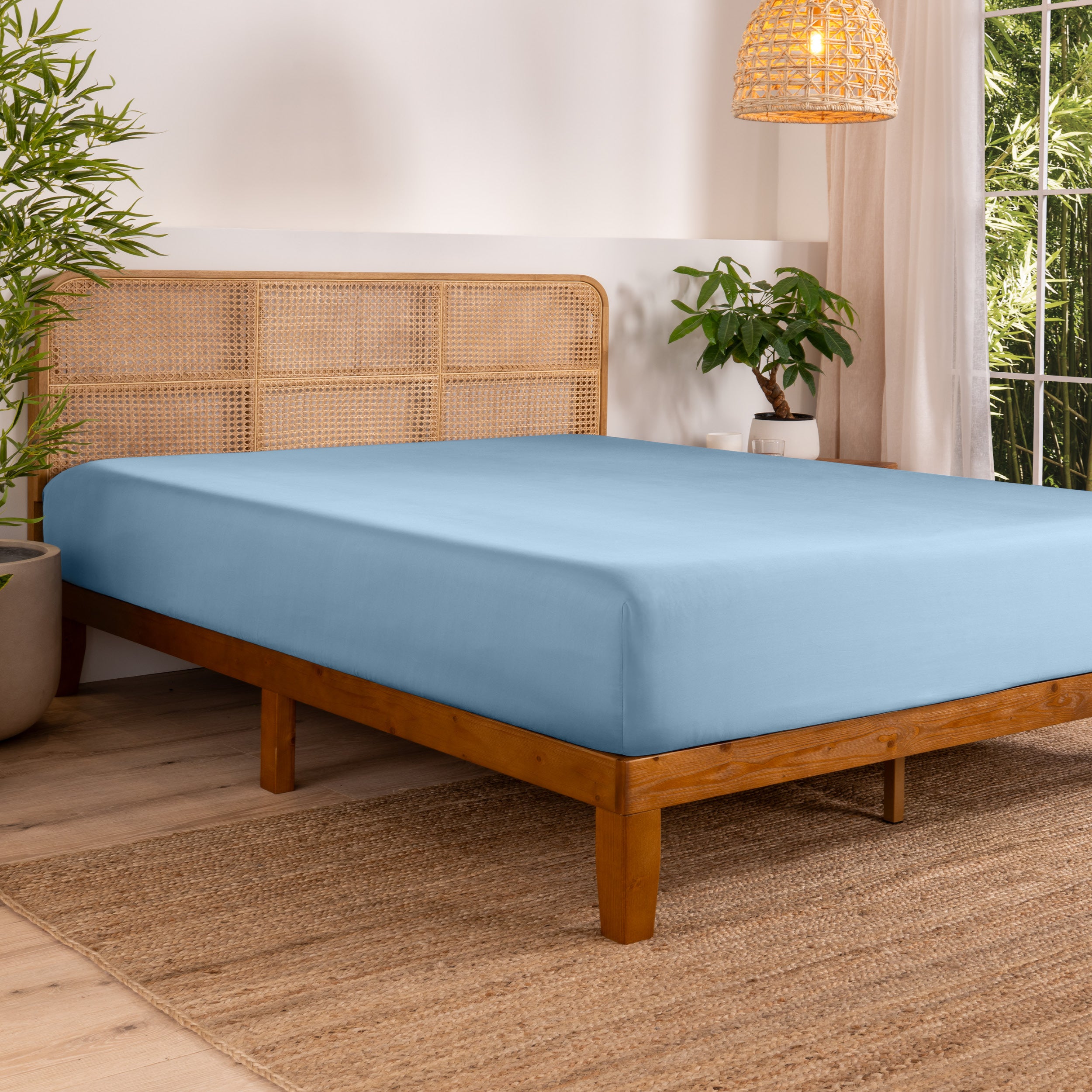 Bed with fitted sheet in a bedroom setting with plants and a lamp.