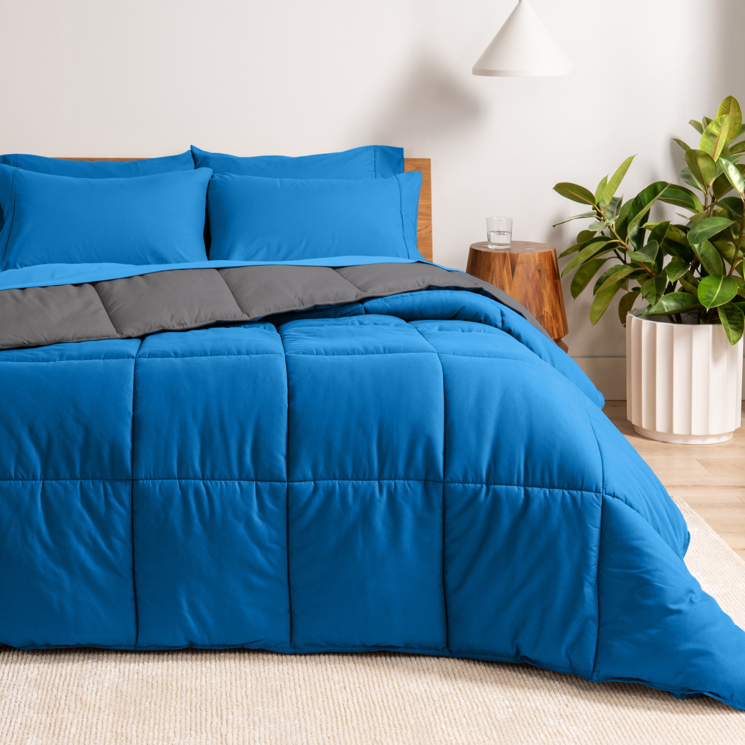 A wooden bed frame with a reversible comforter and sheet set on the mattress in a bright warm room.