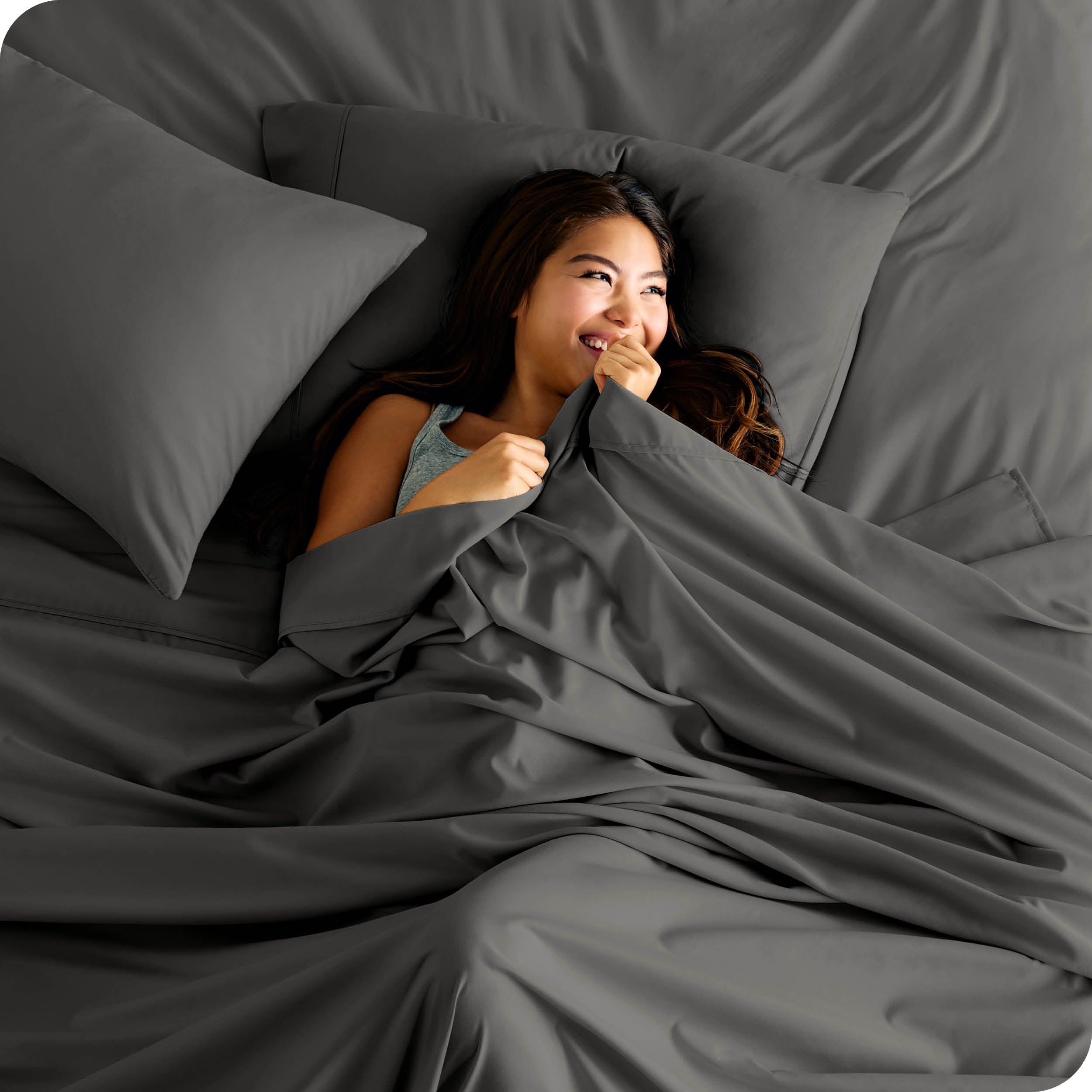 A smiling woman is lying in bed with a top sheet pulled up to her chin