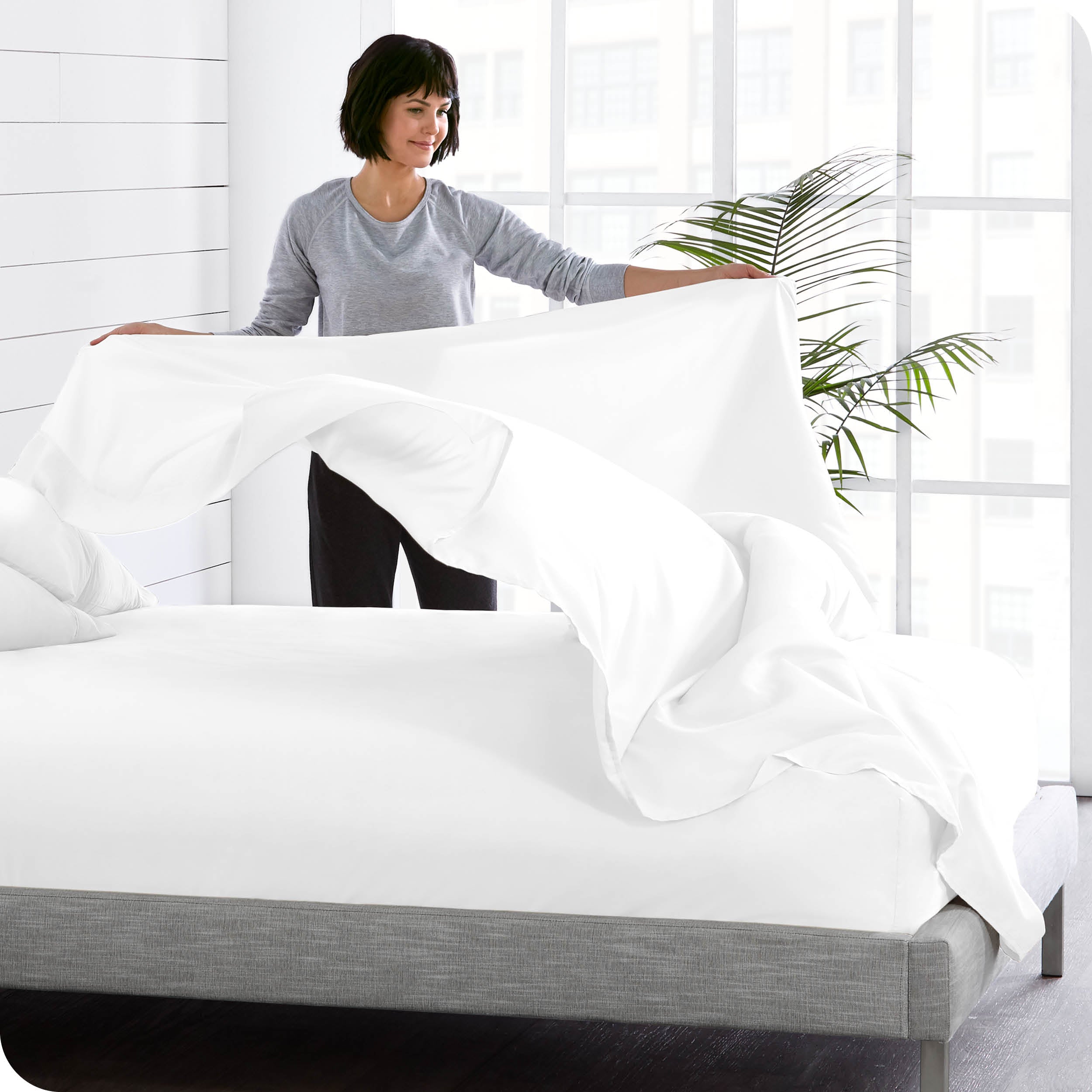 A woman is standing at the side of a bed putting a flat sheet on top of the mattress which has a fitted sheet on it